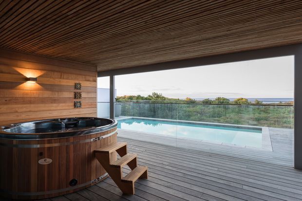 A sophisticated indoor-outdoor spa area with a round timber-clad hot tub in the foreground, set beneath a richly slatted timber ceiling. Timber wall panelling, integrated wall lighting, timber decking, frameless glass balustrading and a rectangular swimming pool create a warm, resort-like setting with a distant coastal view.