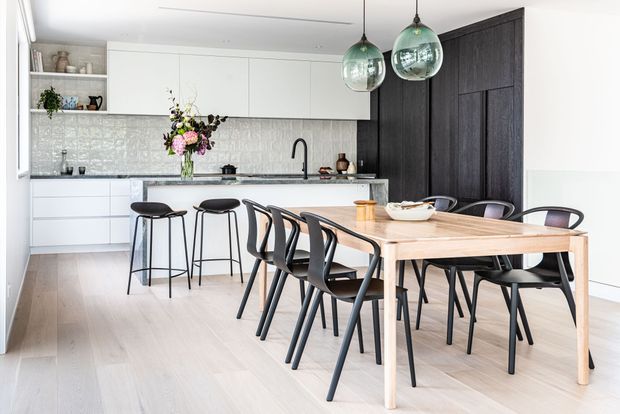 Bright open-plan kitchen and dining area with white handleless cabinetry, a pale textured splashback, dark timber feature cabinetry, a stone island with black tapware, and a light timber dining table surrounded by black chairs. Two green glass pendant lights hang above the island, and pale timber flooring runs throughout.