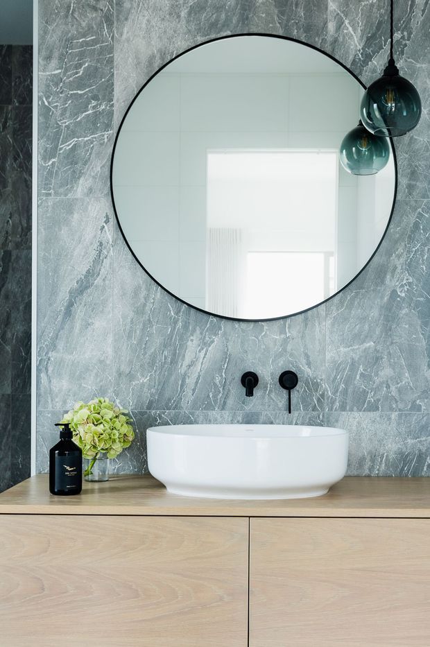 Contemporary bathroom vanity featuring a large round black-framed mirror, grey veined marble-look wall finish, a white vessel basin, matte black wall-mounted tapware, dark glass pendant lights, and light timber cabinetry with a benchtop.