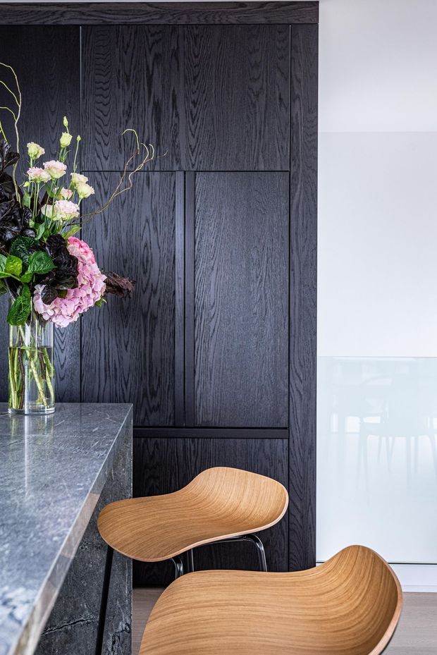 A refined interior vignette featuring a dark, vertically grained timber-paneled wall with integrated joinery, a grey stone island or counter in the foreground, and two sculptural light-wood chairs. A floral arrangement sits in a glass vase on the counter, while a bright glazed area is visible to the right.