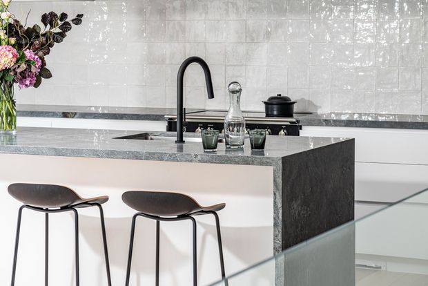 Contemporary kitchen with a central island featuring a dark stone benchtop and waterfall end, integrated sink, matte black gooseneck tap, white cabinetry, and glossy white tiled splashback. Two modern bar stools sit at the island, with subtle decorative styling and strong natural light.