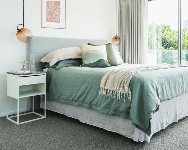 A bright, serene bedroom with a large upholstered bed dressed in layered sage green and cream bedding, a pale grey headboard, white walls, grey carpet, slim white bedside table, globe pendant lights, framed botanical artwork, and full-height curtains beside a large glazed opening.