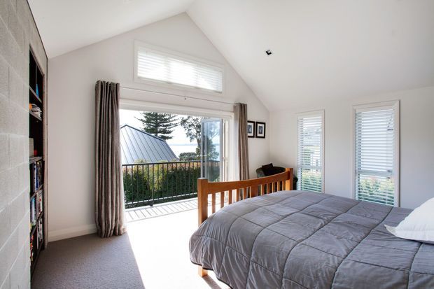 Bright contemporary bedroom with a pitched, partly vaulted ceiling, grey carpet, white walls, and a wooden bed with grey quilt. Full-height glazing and a glazed door open to a small balcony, with white plantation shutters and striped curtains. Built-in open shelving is visible along the left wall.