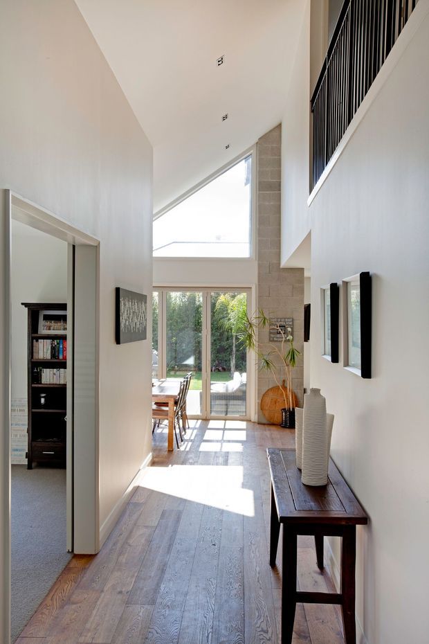A bright, narrow interior hallway with a double-height void and upper-level black metal balustrade. The corridor features wide timber flooring, white painted walls, a dark timber console table with decorative vases, framed wall art, and a large glazed opening at the rear that brings in sunlight and views of greenery. A carpeted room with built-in shelving is visible through an opening on the left, while a tall pale masonry feature wall rises near the rear glazing.