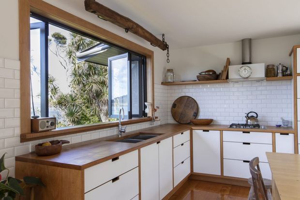 Bright, warm kitchen with white shaker-style cabinetry, timber benchtops, white subway-tile splashback, and a large timber-framed casement window overlooking greenery. Open timber shelving, exposed rustic beam detailing, and warm wood flooring add a relaxed farmhouse character.