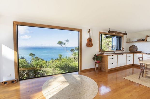 Bright open-plan coastal living and kitchen area with extensive ocean views through a large timber-framed window and a smaller kitchen window. The room features polished timber flooring, white walls and cabinetry, timber benchtops, open shelving, a dining table, round woven rug, and a wall-mounted ukulele. Lush vegetation and water are visible outside.