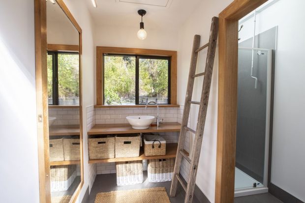 A bright, compact bathroom with a timber-framed window, white tiled walls, a timber vanity with open shelving and woven baskets, and a white vessel basin. A rustic timber ladder leans against the wall, while a glass shower enclosure is visible at right. A large mirror reflects the vanity and window, enhancing the sense of space.