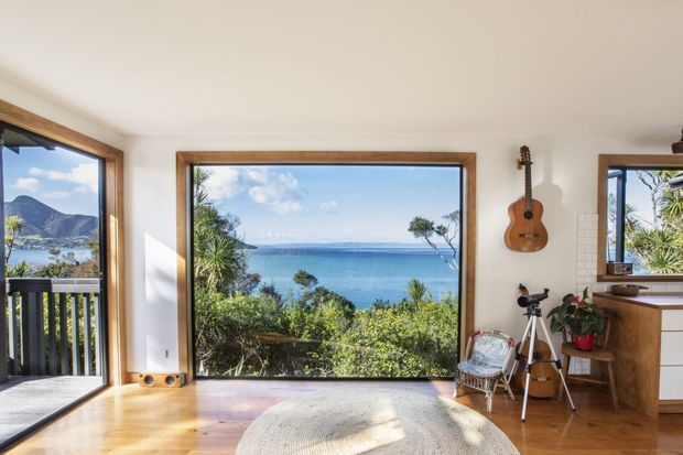 Bright coastal interior with warm timber flooring and timber-framed floor-to-ceiling glazing overlooking a bay and surrounding landscape. A small guitar, telescope, chair, rug, and timber cabinetry add a relaxed residential character.