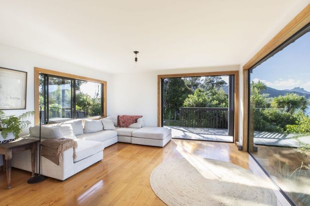 Bright contemporary living room with a large L-shaped sectional sofa, warm timber flooring, white walls, and extensive timber-framed glazing opening onto a deck and leafy mountain landscape. Natural light, indoor plants, and neutral furnishings create an airy, relaxed atmosphere.