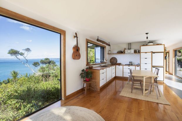 Bright open-plan kitchen and dining area with warm timber flooring, white cabinetry, timber-framed windows, and extensive glazing overlooking a coastal landscape. The kitchen includes a compact island or peninsula, tiled splashback, open shelving, and wood-toned benchtops, with a small dining table nearby. Natural light and indoor plants create an airy, relaxed atmosphere.