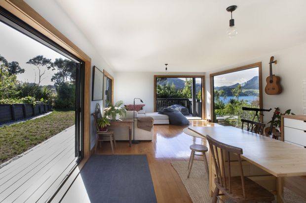 Bright open-plan living and dining space with extensive timber-framed glazing opening onto a deck and landscaped garden. The room features polished timber flooring, a large light-wood dining table, neutral upholstered seating, a compact living area, simple pendant lighting, and a wall-mounted acoustic guitar. A scenic wooded and mountainous landscape is visible through the windows.