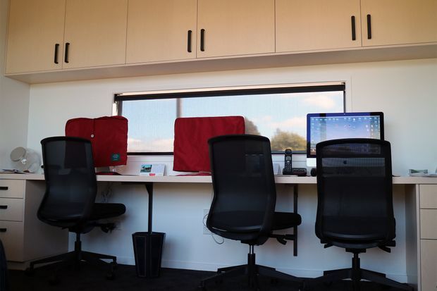 Compact shared home office with three black ergonomic office chairs arranged along a long built-in desk beneath a wide window. The room has pale walls, light timber overhead cabinetry with black handles, white desk surfaces and drawers, computer equipment, and red fabric panels or coverings behind the workstations.