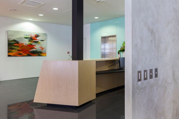Contemporary reception area with a sculptural timber reception desk, dark tiled flooring, pale textured walls, a black structural column, recessed ceiling lighting, and a colourful abstract artwork. A mirrored or glazed wall reflects the reception zone and an elevator lobby beyond.