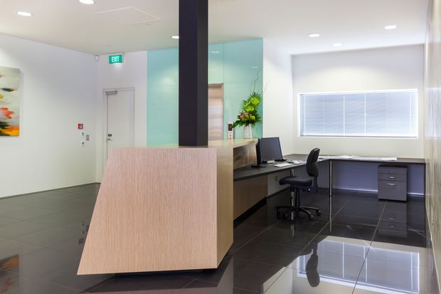 A bright contemporary commercial reception and office area with a light timber reception desk in the foreground, dark glossy tiled flooring, built-in workstations, computers, office chairs, and a large window with blinds. A pale turquoise glass or glazed feature wall and black structural column add contrast, while artwork and an exit door are visible in the background.