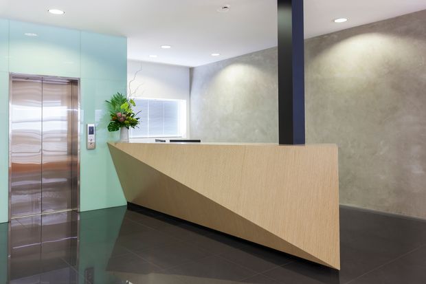 Modern commercial reception lobby with a sculptural light-wood reception desk, dark tiled floor, textured grey feature wall, glossy stainless-steel elevator doors, pale aqua wall panels, and a dark vertical structural column. Recessed ceiling lighting and a floral arrangement complete the polished reception setting.