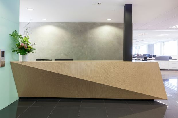 Modern commercial reception area featuring a sculptural light-wood reception desk with a dramatic angular front, dark polished floor tiles, a textured grey feature wall, and a black vertical column. An office workspace is visible beyond the reception desk, with a floral arrangement adding colour at the side.