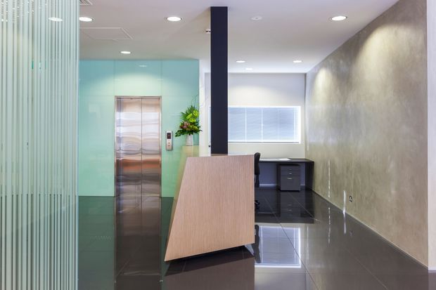 Contemporary commercial reception and office interior with a sculptural light-wood reception desk, glossy dark tiled flooring, a textured grey feature wall, frosted aqua glass around a stainless-steel elevator, and a compact workstation at the rear. The space is brightly lit with recessed ceiling lights and horizontal blinds.