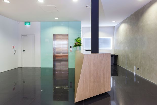 A contemporary commercial reception or lift lobby with a highly reflective dark tiled floor, pale walls, a textured feature wall, stainless-steel elevator doors set within a turquoise glass surround, and a timber reception desk beneath a dark structural column. Bright recessed ceiling lighting and glazed windows create a clean, polished atmosphere.