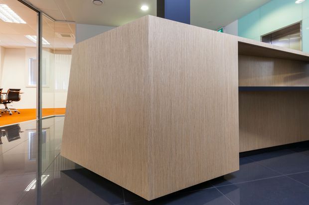 Modern commercial interior featuring a large light wood-grain reception or storage counter with open shelving, dark tiled flooring, glass partitions, and a bright orange-accented office area visible beyond.