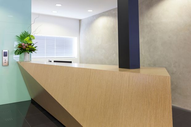 A contemporary reception area featuring a large angular light-wood reception desk with a white benchtop, dark tiled flooring, a textured grey feature wall, and a bold dark structural column. A frosted glass partition, window blinds, recessed ceiling lights, and an intercom/access panel complete the clean commercial interior.