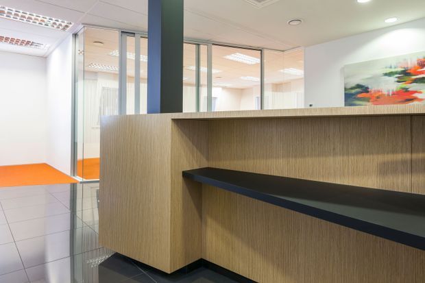 A contemporary commercial reception area with a large built-in timber-look reception counter and dark inset work ledge. Full-height glazed partitions reveal bright office spaces beyond, while orange carpet provides a vivid accent against grey tiled flooring. Recessed ceiling lighting and abstract wall artwork complete the professional fit-out.
