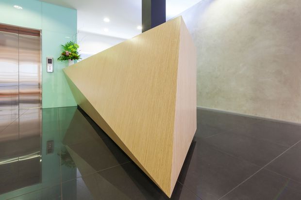 Contemporary commercial or residential lobby featuring a sculptural light-wood reception desk or partition with a sharply angled geometric form. The space has glossy dark floor tiles, a pale textured wall, a turquoise glass wall, recessed ceiling lights, and a stainless-steel elevator.