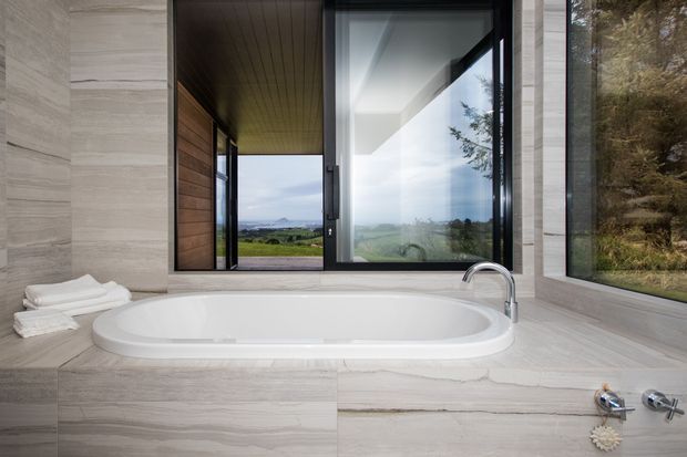Contemporary bathroom featuring a freestanding white bathtub set into a pale stone-tiled surround, with matching stone-look wall finishes, dark timber ceiling, and expansive black-framed glazing overlooking a rural landscape.