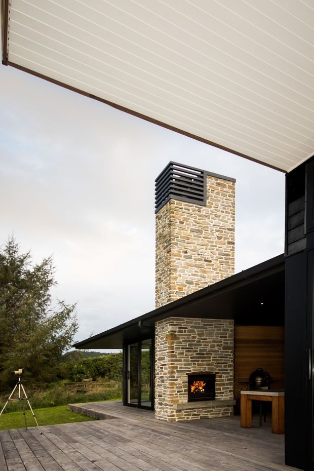 A covered outdoor terrace beside a contemporary dark-clad house, featuring a tall natural-stone chimney with a black louvred cap and an integrated open fireplace. Timber decking extends across the foreground, with a partially enclosed outdoor seating or cooking area visible to the right. The architecture has a strong linear roof canopy, black cladding, expansive glazing, and a rural landscaped setting.
