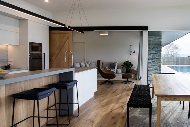 Open-plan contemporary kitchen and living area with a white island, timber-look cabinetry, dark bar stools, warm timber flooring, and a relaxed seating zone. Black linear pendant lighting spans the kitchen, while large glazing connects the dining area to an outdoor swimming pool and landscaped setting.