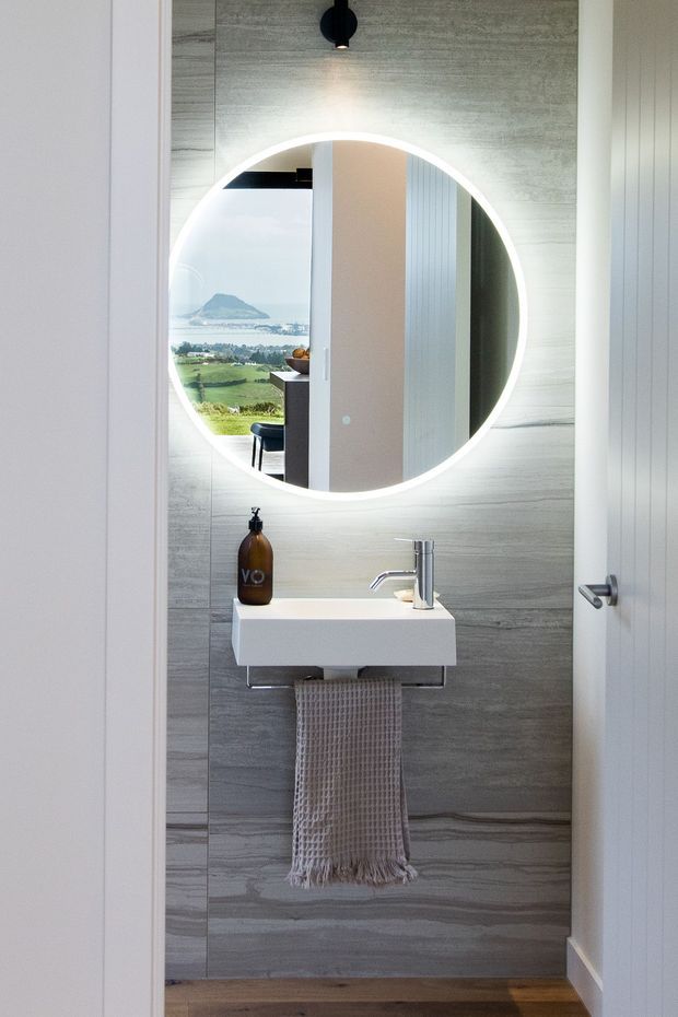 A compact, contemporary bathroom or powder room viewed through an open doorway. The room features a floating white rectangular basin with a chrome tap, a large round backlit mirror, grey wood-look or stone-effect wall tiling, a small black pendant light, and a textured neutral hand towel. The mirror reflects a bright coastal landscape and part of an adjacent interior, but the primary subject is the bathroom vanity area.