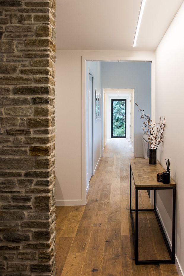 A bright, narrow residential hallway with warm timber flooring, white walls and trim, a long linear ceiling light, and a slim console table with black metal framing. A prominent exposed rough stone-brick column defines the left side, while a glazed door at the far end provides a view to greenery outside.