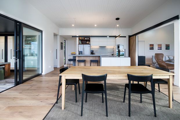 A bright open-plan dining and kitchen area with a large timber dining table and black chairs in the foreground. The space features light timber flooring, white walls and a white paneled ceiling, a contemporary white kitchen with a stone-look island benchtop, black-framed glazed doors to the left, and a partially visible lounge area to the right.
