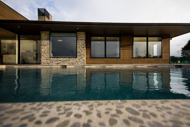 Exterior view of a low, single-storey modern residence seen across a swimming pool. The facade combines horizontal timber cladding, dark-framed floor-to-ceiling glazing, a broad flat roof, and a central natural stone-clad chimney or feature wall. The pool and textured pebble-finish paving dominate the foreground.