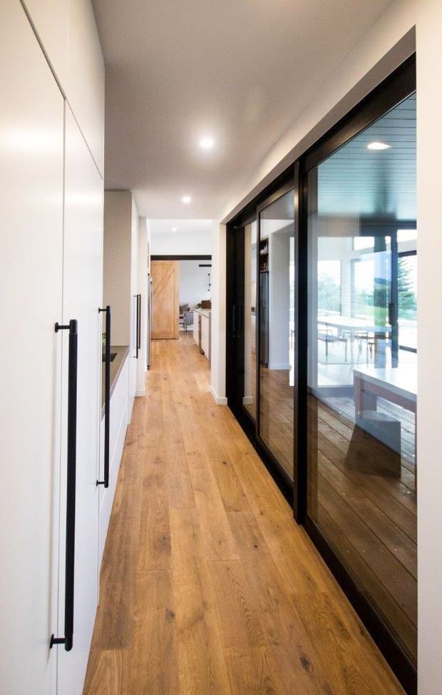 A bright, narrow residential hallway with warm timber flooring, tall white built-in cabinetry and long black handles along the left wall. Full-height black-framed glazed doors on the right open visually onto a light-filled living or dining area. Recessed ceiling lights and clean-lined finishes create a contemporary, minimalist feel.