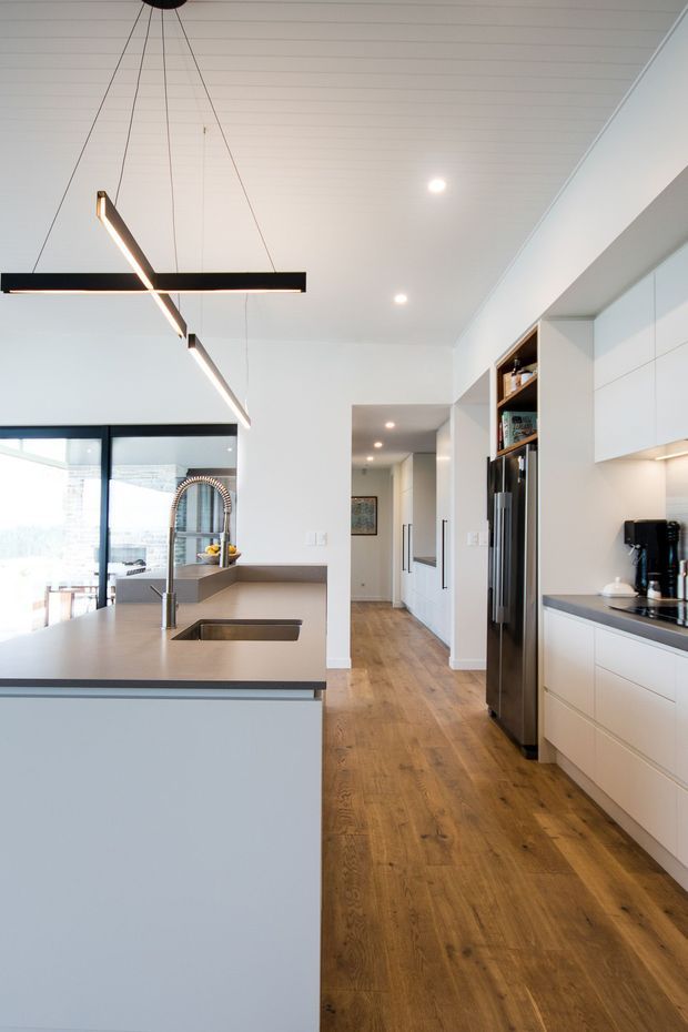 Bright contemporary kitchen with a large island and integrated sink, white flat-panel cabinetry, dark stone-look benchtops, timber flooring, stainless steel appliances, and a striking linear black pendant light. Glazed doors and an arched feature window bring in natural light, while a hallway extends beyond the kitchen.