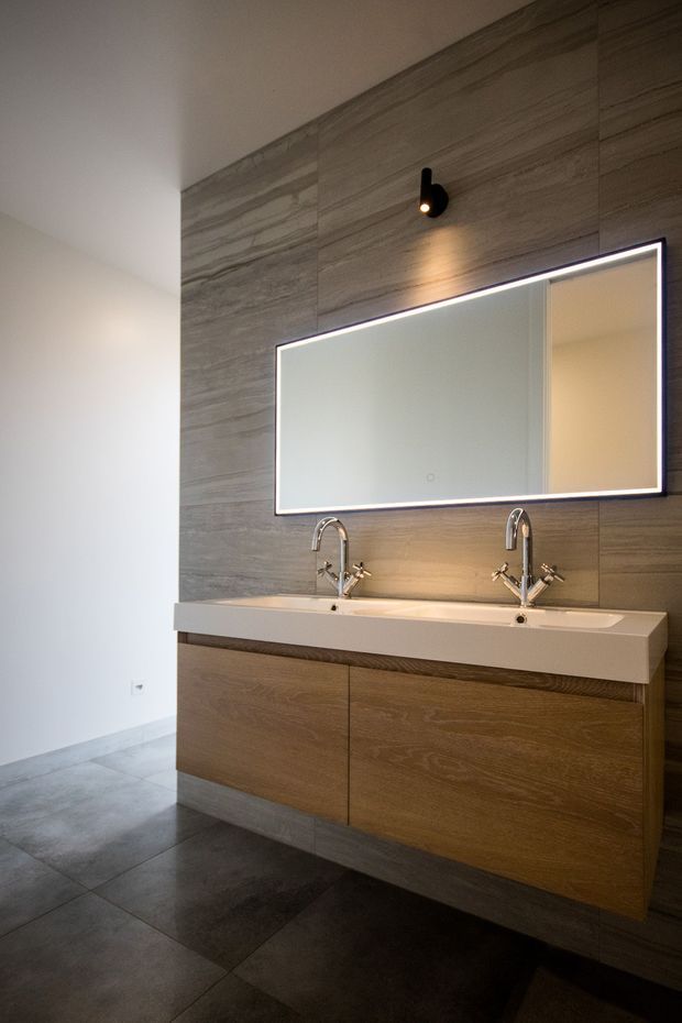 Contemporary bathroom featuring a wall-mounted double vanity with timber-look cabinetry, a white integrated benchtop with twin basins, chrome tapware, a large illuminated mirror, and grey stone-look wall and floor tiling. A black wall spotlight provides focused lighting above the vanity.