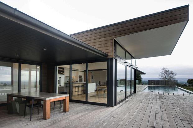 Contemporary coastal residence viewed from a broad timber deck, with expansive floor-to-ceiling glazing, dark timber-clad roof overhangs, and a cantilevered upper roof form. The open-plan interior is visible through the glazing, while the deck leads toward a rectangular swimming pool and distant coastal landscape.