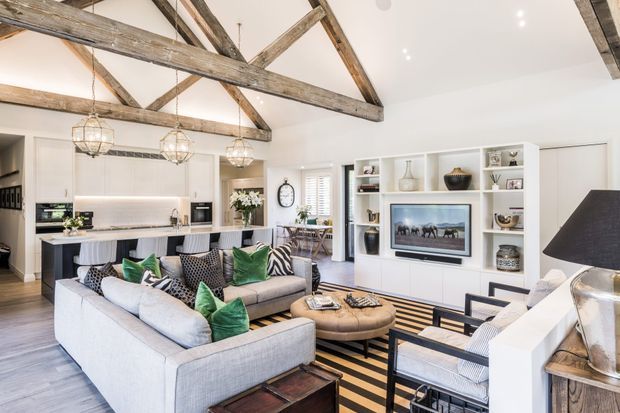 Bright open-plan living room with a large sectional sofa, armchairs, built-in media and display cabinetry, and an adjoining white kitchen with island seating. The room features a high pitched ceiling with exposed rustic timber trusses, white walls and joinery, patterned furnishings, layered rugs, and pendant lighting.