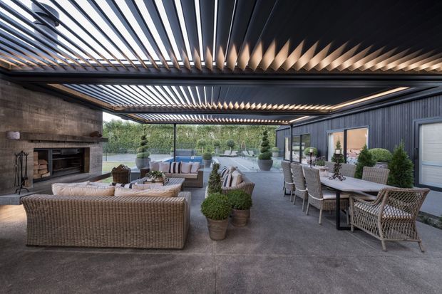 A spacious covered outdoor entertaining area with a dark metal louvred pergola roof, concrete flooring, a large masonry fireplace, woven outdoor sofas and armchairs, and a dining table beside a dark cladded building. The space opens onto landscaped grounds and a pool or water feature in the background.