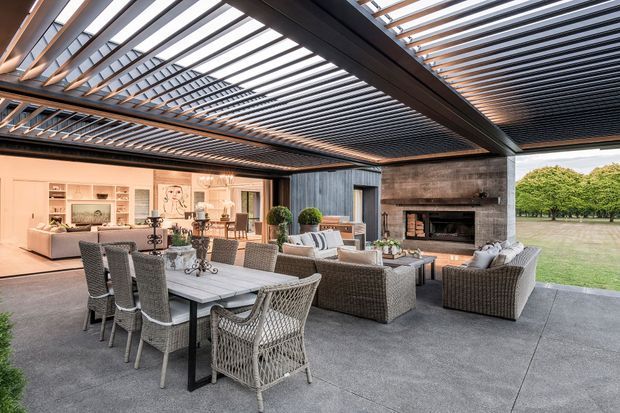 A sophisticated covered outdoor entertaining area with a large dining table, woven dining chairs, lounge seating, and a built-in fireplace. A retractable or louvred overhead structure creates a dramatic slatted ceiling, while the space opens onto a lawn and landscaped garden. The adjoining interior living area is visible through the wide opening.