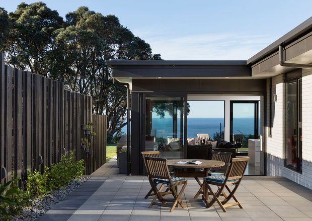 A contemporary coastal home's paved outdoor terrace overlooking the ocean. The courtyard-like patio features a timber outdoor dining set, dark-framed floor-to-ceiling glazing, a low flat roof, light brick walling, and a tall dark timber privacy fence with landscaped planting along the side.