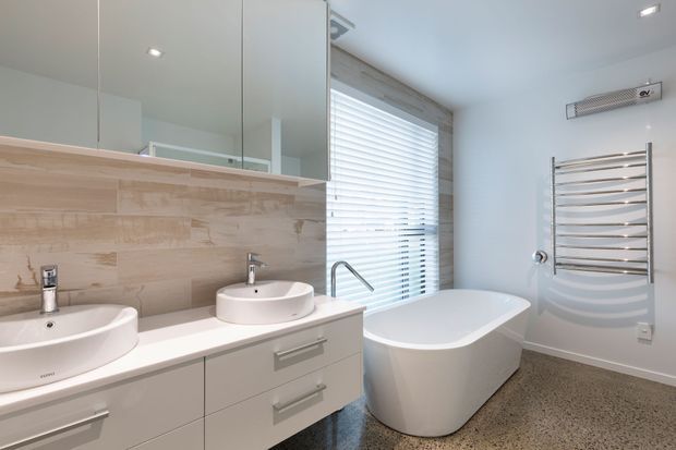 Bright contemporary bathroom with a freestanding white bathtub beside a large window, double vessel basins on a pale built-in vanity, a mirrored wall cabinet, beige stone-look tiled splashback, heated towel rail, and terrazzo-style flooring. The room has clean lines, recessed ceiling lighting, and a neutral palette.