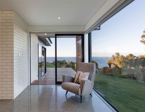 A bright contemporary living area or sunroom with extensive floor-to-ceiling glazing overlooking a coastal landscape and ocean. The room features a polished speckled floor, white painted brick walls, a white panelled ceiling with recessed lighting, dark-framed sliding glass doors, and a striped upholstered lounge chair. A timber deck and lawn are visible outside.