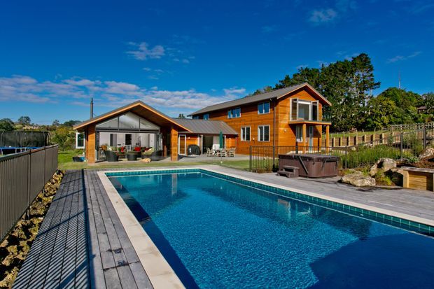 Exterior view of a multi-level timber-clad house with a glazed gable-roofed pavilion, expansive swimming pool, and surrounding timber decking. The property has a warm natural wood facade, dark metal roofing, large windows, and a covered outdoor entertaining area, set in a landscaped suburban or semi-rural environment under a bright blue sky.