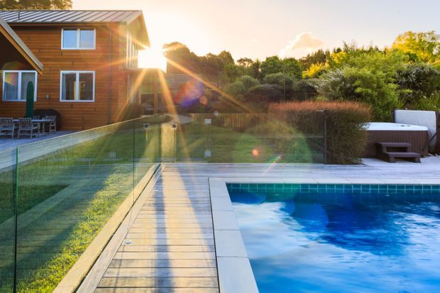 Exterior view of a contemporary timber-clad two-storey house opening onto a landscaped backyard with a swimming pool. A glass balustrade separates the timber pool deck and walkway from the lawn, while outdoor seating is visible beside the pool. Warm late-afternoon sunlight creates a bright, relaxed residential atmosphere.