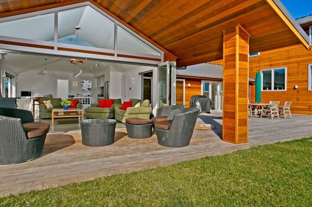 A spacious covered outdoor deck and entertaining area adjoining an open-plan living room. The deck has timber flooring, a broad timber-lined soffit, exposed structural posts, outdoor lounge furniture, and a dining setting. Large folding or sliding glazed doors open the living space directly onto the deck, while the adjoining house features warm horizontal timber cladding and a high glazed gable.