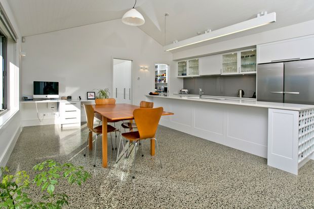 Bright open-plan kitchen and dining area with a long white kitchen featuring integrated cabinetry, glazed upper cupboards, a grey backsplash, stainless-steel appliances and a peninsula. A timber dining table with orange upholstered chairs sits centrally, while a small living area and large window are visible to the left. The room has a polished speckled terrazzo-style floor, white walls and ceiling, and contemporary linear lighting.