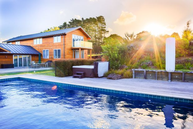 Exterior view of a multi-storey timber-clad home beside a swimming pool. The property features a blue pitched metal roof, extensive glazing, a covered outdoor dining area, timber pool decking, raised garden beds, and lush landscaped planting, photographed in warm late-afternoon sunlight.
