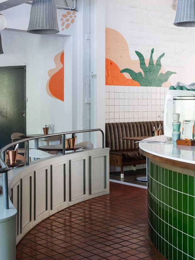 A stylish café interior with a curved green tiled service counter, curved light-grey paneled cabinetry, dark brown tiled flooring, upholstered booth seating, and a small round tiled table. White-painted brick walls feature bold abstract orange and green botanical murals, while woven pendant lights add texture overhead.