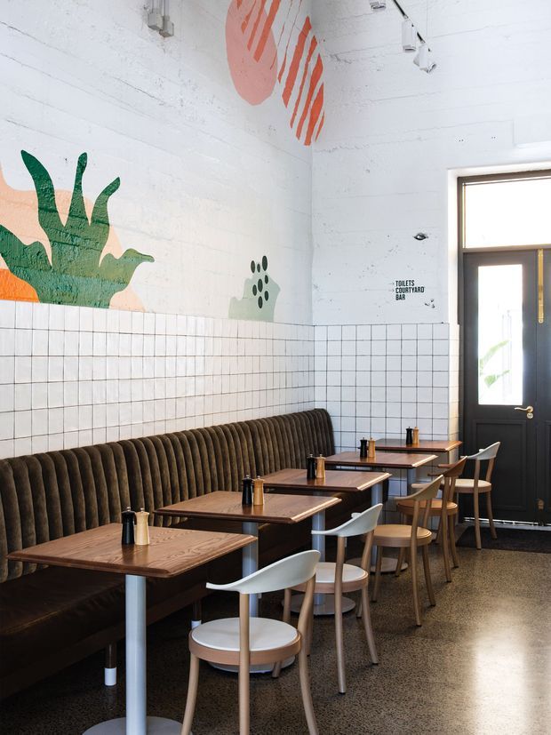 Bright contemporary café or casual dining interior with a long ribbed upholstered banquette, multiple timber dining tables, mixed timber and white chairs, polished dark flooring, white tiled lower walls, and white painted upper walls featuring large colourful abstract botanical murals. A dark framed glazed door and directional signage are visible at the end of the room.